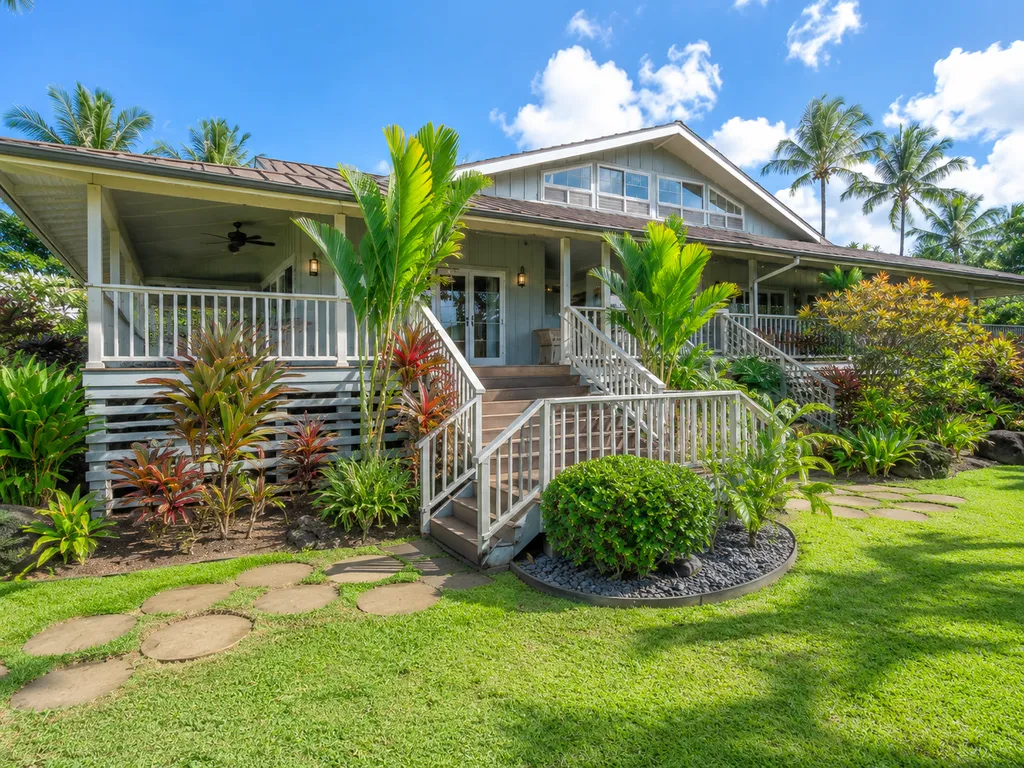 A white house with a porch, surrounded by lush greenery and a clear blue sky.
